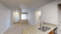 This single-floor space showcases an open-concept design that seamlessly connects the kitchen and living areas. The kitchen features granite countertops with subtle grey and beige veining, paired with a stainless steel double-basin sink and a modern pull-down faucet. The countertop extends as a peninsula, creating a natural visual boundary between the kitchen and living space.

The living area is finished with neutral-toned carpet and warm beige walls, creating a cohesive and welcoming atmosphere throughout. A ceiling fan with integrated lighting is centrally positioned, providing both air circulation and ambient light. A window fitted with white horizontal blinds allows natural light to filter into the space, illuminating the front entryway, which features a white paneled door.

An electrical panel is neatly recessed into the wall, and a textured popcorn ceiling runs throughout. HVAC vents are strategically placed for efficient climate control. The open layout and neutral color palette give the space a clean, functional character well-suited for everyday living.