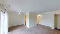This single-floor space features a spacious, open living area finished with wall-to-wall neutral-toned carpeting that adds warmth and comfort underfoot. The room is defined by clean white walls and a smooth ceiling equipped with a ceiling fan and light fixture, providing both air circulation and ambient lighting throughout the space.

A partial dividing wall creates a natural separation within the layout, subtly defining different functional zones while maintaining an open feel. Through an opening in this partition, the kitchen is visible, showcasing warm wood-toned cabinetry that introduces a natural contrast against the neutral color palette of the surrounding walls. The kitchen appears well-appointed with upper and lower cabinetry, hinting at ample storage.

An additional area extends to the right side of the living space, illuminated by a wall-mounted light, suggesting a secondary zone that could serve various purposes. Multiple electrical outlets are positioned along the baseboards throughout, offering practical convenience. The overall ambiance is clean, bright, and functional, with a simple and understated design that provides a versatile foundation for personalization.
