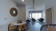 This single-floor space showcases a thoughtfully designed open-plan layout that seamlessly connects the dining and living areas. The interior is defined by its industrial-chic aesthetic, highlighted by exposed ductwork and piping running along the ceiling, which contrasts elegantly with the clean white walls and smooth, polished concrete floors throughout.

The dining area features a round marble-top table paired with woven rattan chairs, adding a warm, organic texture to the space. A striking sunburst mirror mounted on the wall serves as a decorative focal point, enhancing the room's visual interest. Adjacent to the dining area, the living space is furnished with dark leather seating arranged on a light area rug, creating a comfortable and cohesive lounge environment.

Large sliding glass doors at the far end of the space flood the interior with natural light while providing direct access to an outdoor area, contributing to an airy, open ambiance. The elevated ceiling height adds a sense of volume and grandeur to the overall layout. Recessed lighting supplements the natural light, ensuring the space remains well-lit throughout. The overall atmosphere is contemporary, functional, and stylish.