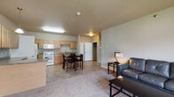 Located at 906 Park Avenue West in Watford City, North Dakota, Unit 209 is a thoughtfully designed single-floor residence that offers a comfortable and functional layout. The open-concept kitchen and dining area form the heart of the unit, featuring light wood cabinetry, granite-style countertops, tile flooring, and a full suite of white appliances including a range, microwave, refrigerator, and dishwasher. A dining table positioned adjacent to the kitchen makes the space ideal for everyday meals. The living room is generously sized, carpeted, and filled with natural light from large windows that overlook the surrounding building exterior. The space comfortably accommodates a full seating arrangement, lending a relaxed and welcoming atmosphere. The bedroom is a standout feature, offering plush carpet, a walk-in closet with built-in shelving, and a sliding glass door that opens to a private balcony with outdoor views. A second bedroom is also present, accessible from the main living area and equally well-lit with exterior-facing windows. The unit's neutral color palette of warm grays and taupes creates a cohesive, calm ambiance throughout, making it a practical and inviting place to call home.