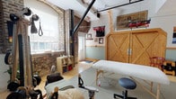 The Palates Room is a thoughtfully designed wellness studio that blends historic architectural character with a comprehensive range of movement and bodywork equipment. Exposed brick walls and warm hardwood flooring establish an inviting, loft-style atmosphere, while white-painted upper walls and arched windows bring in natural light, enhancing the open, airy feel of the space.

The studio is outfitted with a padded massage/treatment table, a wooden ladder barrel and wall unit, a reformer-style apparatus, overhead suspension equipment, and aerial straps and rings suspended from ceiling-mounted tracks. Interlocking floor mats define the primary movement area, providing a safe, cushioned surface for floor-based work. A large wall mirror amplifies the sense of space and allows practitioners and clients to monitor alignment and form.

Organizational elements include open shelving units stocked with folded linens, yoga mats, bolsters, and accessories, as well as a substantial wooden barn-style cabinet for additional storage. A sheer curtain partition offers a flexible way to section the space for privacy when needed. Decorative artwork and a small collection of singing bowls add a serene, holistic ambiance. Exposed HVAC ductwork and ceiling-mounted track lighting contribute to the studio's functional yet characterful industrial aesthetic.