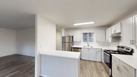 This single-floor residence offers a clean, modern aesthetic throughout its well-organized layout. The kitchen is a standout feature, outfitted with white shaker-style cabinetry, light countertops, and a full suite of stainless steel appliances including a refrigerator, dishwasher, and electric range with an overhead vent hood. A built-in peninsula provides additional counter space and separates the kitchen from the adjoining living area. Wood-look luxury vinyl flooring runs through the main living spaces, adding warmth and durability. The living area is bright and open, featuring a ceiling fan with light fixture, a window with blinds, and sliding glass doors that lead directly to an outdoor patio with views of a lush, landscaped backyard. A second open living or dining space also features sliding glass doors, allowing natural light to fill the room. The bedrooms are carpeted and each include baseboard heating and windows fitted with blinds, ensuring comfort and privacy. The bathroom is finished with a wide vanity, large wall mirror, a bright vanity light fixture, and a walk-in shower with a glass sliding door. The overall palette of crisp whites, neutral tones, and natural textures creates a cohesive and inviting atmosphere throughout the home.