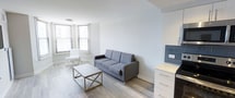 This single-floor space offers a clean and contemporary living environment defined by its minimalist aesthetic and thoughtful design. The main living area is bathed in natural light, courtesy of multiple windows fitted with horizontal blinds that allow for adjustable brightness throughout the day. Smooth, light-toned hardwood flooring runs throughout, complementing the crisp white walls and creating a warm yet modern atmosphere. The living space is well-proportioned, providing a comfortable setting for both relaxation and everyday living. A sleek gray sofa serves as the centerpiece of the room, paired with a minimalist gold-accented coffee table and a white accent chair positioned near the bay-style window corner — an ideal spot for reading or unwinding. A white dresser with gold hardware provides functional storage while contributing to the room's cohesive, contemporary palette. The overall ambiance is light, airy, and uncluttered, with a neutral color scheme that lends itself well to a variety of interior styling preferences. High, smooth ceilings and clean architectural lines reinforce the modern character of the space, making it feel open and inviting throughout.