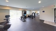 Located at 6501 Hil Mar Drive in District Heights, Maryland, this fitness center offers a comprehensive workout facility spread across a single floor. The space is thoughtfully organized into distinct zones, each designed to support a variety of exercise routines and fitness goals.

The cardio area is outfitted with multiple treadmills and elliptical machines, complemented by wall-mounted flat-screen televisions for entertainment during workouts. Large windows fitted with blinds allow ample natural light to fill the space, creating a bright and energizing atmosphere. Full-length mirrors are strategically placed throughout to assist with form and to enhance the sense of openness.

Adjacent areas accommodate a range of strength-training equipment, including cable machines, shoulder press stations, and a multi-function power tower. Recessed lighting provides consistent, even illumination throughout the facility. The durable rubber flooring throughout the space offers both safety and comfort underfoot during all types of exercise.

A vending machine is available on-site for added convenience. The neutral, light-colored walls and clean layout contribute to a functional and welcoming environment. Multiple windows across different sections of the facility frame views of the surrounding greenery, adding a refreshing visual element to the workout experience. This fitness center is well-suited to serve a broad range of fitness needs in a practical and accessible setting.
