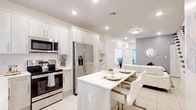This two-story home showcases a cohesive modern design defined by a crisp, all-white palette, recessed lighting, and large-format porcelain tile flooring throughout the main level. The kitchen is a standout feature, outfitted with floor-to-ceiling light wood cabinetry, stainless steel appliances including a French door refrigerator, electric range, and built-in microwave. A central island with a white quartz countertop doubles as a dining surface, accommodating seating for casual meals. The kitchen flows seamlessly into the open-concept living area, which features a built-in shelving niche and natural light filtering through roller-shaded windows. A staircase with dark wood treads and white balusters connects the main level to the upper floor, with a full bathroom and closet storage visible nearby. The bedrooms vary in character: one features an all-white finish with tile flooring and a window with a roller shade, while another offers warm wood-toned furniture and dual windows. A full bathroom includes a tub-shower combination, wood-finish vanity, and white quartz countertop. A dedicated laundry area houses a full-size top-load washer and dryer. The overall ambiance is clean, bright, and contemporary.
