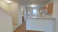 This 2-floor space is designed with a practical, open layout that connects the main living and kitchen areas seamlessly. The kitchen is equipped with medium-toned wood cabinetry, a white electric range, a refrigerator with built-in microwave overhead, and a peninsula counter that doubles as a breakfast bar, providing additional workspace and casual seating. The countertops feature a light, neutral finish that complements the overall color scheme.

The flooring throughout the main level consists of hardwood-style planks in a warm, light tone, while a carpeted area is visible near the staircase, which transitions to the second floor. The walls are finished in a clean, bright white, contributing to an airy and open atmosphere. Natural light filters in through a window near the front entry door, brightening the space.

A hallway leads to a bathroom, partially visible through an open door, and a front entry door with a decorative glass panel allows additional light into the space. Ceiling-mounted lighting provides consistent illumination throughout. The overall layout is efficient and straightforward, offering a well-connected flow between living, dining, and kitchen areas.