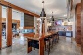 Located at 837 NW Donovan Rd in Lee's Summit, Missouri, this single-floor community lobby space blends industrial and modern design elements into a cohesive, welcoming environment. Polished concrete floors extend throughout, reflecting light and lending an open, contemporary feel to the entire area.

A centerpiece long bar-height communal table with a rich wood-toned surface is surrounded by metal and wood bar stools, creating a practical gathering and workspace. Overhead, a series of bold, tiered cylindrical pendant lights add dramatic visual interest while providing functional illumination. Exposed silver ductwork traces the ceiling, reinforcing the space's industrial aesthetic.

The service station along the back wall features dark cabinetry, a textured tile backsplash, a mounted flat-screen display, and a coffee and refreshment bar stocked for resident convenience. Gooseneck wall sconces accent this area with a nod to vintage industrial style.

Wood-clad structural columns with warm, natural grain detailing define zones throughout the space and anchor banks of black resident mailboxes set against walls featuring large-scale botanical murals. Barn-style sliding doors with heavy timber hardware separate the lobby from an adjacent lounge area, which is visible through the opening and offers comfortable seating and natural light from exterior windows. Together, these elements create a functional, stylish, and inviting community hub.
