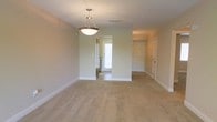 This single-story space presents an open, versatile layout bathed in neutral tones that create a calm, welcoming atmosphere. The main living area features wall-to-wall beige carpeting that adds warmth and comfort underfoot. A centrally hung bowl-style pendant light serves as a subtle focal point, providing functional overhead illumination while complementing the understated aesthetic of the space.

The layout is defined by a partial wall or column division that separates the main living area from an adjacent space, which benefits from natural light filtering through a window fitted with blinds. This segmentation adds architectural interest while maintaining an open, connected feel throughout.

White baseboards run along the perimeter of the walls, providing crisp contrast against the light green-toned walls. A white paneled door is visible along the hallway corridor, indicating additional rooms or storage beyond the main living area. The ceiling is smooth and standard height, with a visible HVAC vent ensuring climate comfort.

Overall, the space is clean, move-in ready, and offers a neutral canvas well-suited for a variety of furnishing styles and functional uses.