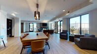 This well-appointed single-floor community space offers a thoughtfully designed layout that seamlessly blends social, dining, and relaxation areas. Light wood-look flooring flows throughout, tying the open-concept zones together cohesively.

The main lounge area is anchored by a striking floor-to-ceiling stacked stone fireplace, creating a warm focal point flanked by comfortable seating arrangements. Large floor-to-ceiling windows and glass doors flood the space with natural light and provide direct access to an outdoor terrace, extending the living experience beyond the interior.

A generously sized dining area features a solid wood table with cognac leather chairs beneath an architectural pendant light, ideal for group gatherings. Adjacent to this is a fully equipped kitchen outfitted with dark wood cabinetry, subway tile and navy mosaic tile backsplash, stainless steel appliances, open shelving, and a large white quartz island with bar seating illuminated by globe pendant lights.

A distinct secondary zone, framed by white columns and deep navy accent walls, leads into a private meeting or club room furnished with a chandelier and dedicated seating. Curated artwork and tasteful décor are displayed throughout, contributing to the space's polished, contemporary ambiance suited for both casual socializing and productive gatherings.