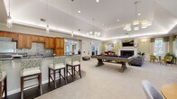 Located at 15 Princeton Dr in Hooksett, New Hampshire, this single-floor community clubhouse offers a thoughtfully designed gathering space with a variety of distinct areas suited for socializing and recreation. The main lounge serves as the heart of the facility, centered around an elegant white-mantled fireplace with slate tile surround, a wall-mounted television, and a tray ceiling with ambient lighting. Comfortable seating, including a plush sofa and upholstered accent chairs, creates an inviting atmosphere complemented by warm neutral tones and large windows that bring in natural light and garden views. Adjacent to the lounge, a billiards area provides recreational space centered around a full-size pool table beneath stylish pendant and chandelier lighting. The fully equipped kitchen features warm wood cabinetry, granite countertops, stainless steel appliances, a central island with bar seating, and tile flooring — ideal for hosting events or casual gatherings. A charming dining nook with built-in bench seating, wainscoting, and an arched window overlooking lush greenery adds an intimate setting for smaller groups. A dedicated sitting room with large windows, abundant natural light, and patterned lounge chairs offers a quiet retreat, while additional flexible space visible in the floor plan accommodates a variety of community activities.