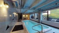 This 2-bedroom, 2-bathroom property includes access to a shared indoor pool facility that serves as a notable amenity. The pool area showcases an industrial aesthetic, characterized by an exposed ceiling with visible ductwork and structural beams painted in cool blue-grey tones, lending the space a modern, urban character. The pool itself is a lap-style design, fitted with stainless steel entry rails and surrounded by tile flooring with rubber mat walkways for safety. Large windows along one wall allow natural light to filter in while offering views of the surrounding outdoor landscape, including what appears to be a wooded hillside. Wall-mounted lighting provides warm ambient illumination throughout the space. Concrete columns frame the perimeter, reinforcing the building's industrial architectural style. An adjacent area visible beyond the pool includes additional amenity space. The overall atmosphere is functional yet inviting, offering residents a convenient indoor aquatic facility regardless of weather conditions. The combination of natural light, industrial detailing, and clean water create a well-balanced recreational environment.