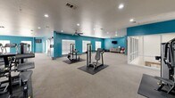 Located at 12025 Paul Eells Dr in North Little Rock, Arkansas, this fitness center offers a single-floor layout with an expansive exercise room designed to accommodate a wide range of workout needs. The space is immediately distinguished by its bold teal accent walls contrasted against neutral gray flooring and a textured white ceiling, creating an energetic yet clean aesthetic throughout.

The facility is organized into clearly defined zones. A dedicated cardio area houses multiple treadmills, elliptical trainers, and stair climbers arranged in rows, allowing ample space between machines for comfortable use. A strength training zone features a robust selection of Cybex and other commercial-grade resistance machines placed on protective rubber mats, catering to both upper and lower body workouts. Free weights, stability balls, and additional functional training equipment are also present.

A glass-partitioned section provides a semi-private area within the larger floor plan, ideal for stretching or focused training. Recessed lighting and multiple ceiling fans throughout ensure the space remains well-lit and comfortable. Windows along the walls allow natural light to supplement the overhead lighting. Multiple entry and exit doors contribute to convenient access and circulation across the facility.