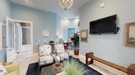 This well-appointed single-floor space serves as both a relaxing lounge and a functional business center, thoughtfully designed with a cohesive coastal aesthetic. The interior is defined by soft powder-blue walls that lend a calm, airy atmosphere throughout, complemented by light marble-style tile flooring and tall ceilings that enhance the sense of openness.

The lounge area is generously furnished with a yellow sofa, coordinating striped armchairs, and patterned accent chairs arranged around a striped area rug and a round coffee table. Decorative touches include potted indoor plants, framed landscape artwork, and ornamental table lamps that contribute warmth and texture to the space. A wall-mounted flat-screen TV is paired with a rustic wood bench, creating a comfortable media viewing area. Striped curtains frame the windows, allowing natural light to filter in while adding visual depth.

Adjacent to the lounge, a dedicated business center features a wood-toned desk, office chair, printer, and computer workstation, making it a practical resource for productivity. The space is topped with an elegant chandelier that adds a refined focal point.

A set of glass-paneled double doors connects to an additional area, further expanding the functionality of this versatile community space. Overall, the design balances comfort and utility with a warm, inviting character.