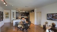 This single-floor residence showcases an open-concept layout that seamlessly connects the kitchen and dining areas, creating a cohesive and functional living environment. The kitchen is outfitted with rich, dark wood cabinetry extending to the upper walls, stainless steel appliances including a refrigerator and microwave, and a breakfast bar with seating that doubles as a casual dining option. Track lighting runs along the ceiling, providing focused illumination across the kitchen workspace. Dark countertops complement the cabinetry, reinforcing a consistent, contemporary aesthetic. A dedicated dining area sits adjacent to the kitchen, furnished with a round glass-top table surrounded by dark wood chairs with cushioned seats. The warm, medium-toned hardwood flooring extends throughout the space, anchoring the room and adding a natural contrast to the darker furnishings. White walls and white-paneled doors provide a clean, bright counterpoint to the bold cabinetry. A decorative wall gallery near the dining area adds a personal touch, while recessed lighting in the ceiling supplements the track fixtures. Two doorways visible in the background suggest additional rooms beyond the main open living area.
