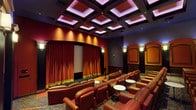 This stunning private home theater is designed to replicate the grandeur of a classic cinema while delivering an intimate, upscale viewing experience. The room is anchored by a large projection screen framed by rich, floor-length velvet curtains in deep red with elegant gold fringe trim, evoking the timeless aesthetic of a traditional movie palace.

Seating is arranged in tiered rows, accommodating a generous number of viewers in plush, cognac-toned leather recliners and matching sofas. Individual seats are generously sized with wide armrests, providing exceptional comfort for extended viewing sessions. Small round side tables are positioned throughout the seating area for added convenience.

The walls are finished in warm crimson and deep burgundy tones, complemented by decorative arched panel columns with ornate gold trim and wall sconces that cast a warm, ambient glow. Dark wainscoting and floor-to-ceiling acoustic drapery panels line the side walls, enhancing both the visual richness and sound quality of the space.

Overhead, a dramatic coffered ceiling features recessed lighting and a soft purple-hued ambient border that adds depth and a theatrical atmosphere. The intricately patterned carpet in jewel tones of red, green, and gold ties the entire design together, completing the cinematic aesthetic of this exceptional entertainment space.