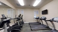 Located at 146 Yester Oaks Dr in Mobile, Alabama, this dedicated fitness center offers a well-appointed, single-floor exercise facility designed to support a well-rounded workout routine. The space is organized into distinct functional zones, providing ample room to move comfortably between equipment.

The cardio area is equipped with multiple TRUE-brand treadmills and elliptical machines, arranged to allow easy access and use. A wall-mounted flat-screen television is positioned for viewing during workouts, and large wall mirrors extend across one side of the room, creating a sense of added depth and allowing users to monitor their form. Natural light filters in through multiple windows fitted with white plantation shutters, complementing the bright overhead fluorescent lighting.

A dedicated strength training area features a two-tier dumbbell rack stocked with a full range of free weights, accompanied by an adjustable workout bench for versatile strength exercises. Motivational framed cycling artwork decorates the walls of this zone, adding a lively, athletic aesthetic. The space is finished with durable patterned carpet tile flooring throughout, and a French-style glass door connects to an adjoining entry area with hardwood flooring, providing a clean and accessible transition into the facility.