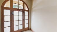 This single-floor space showcases notable architectural character through its carefully preserved classical details. The entrance is defined by elegant French doors framed by warm wood trim and topped with a striking arched transom window, allowing natural light to filter into the entryway. The frosted glass panes of the doors provide privacy while maintaining a sense of openness, and the arched surround adds a refined, traditional aesthetic to the entry point.

Inside, a staircase with dark hardwood treads and a light wood handrail rises prominently, its clean lines and classic balustrade contributing to the overall architectural integrity of the space. The neutral, cream-toned walls throughout provide a clean and versatile backdrop, complemented by warm hardwood flooring that extends along the corridor. A hallway stretches beyond the staircase, lined with doors that suggest multiple rooms or functional areas branching off from the main circulation path. Crown molding and wood baseboard trim add finishing detail throughout. The overall ambiance is one of traditional craftsmanship, with a practical, well-organized interior layout suited to a variety of uses.