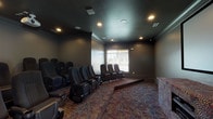 This private theater is designed to replicate a true cinematic experience within a single-floor layout. The room is finished with dark, muted walls and crown molding that contribute to its immersive, light-controlled atmosphere. A ceiling-mounted projector directs content onto a large widescreen projection display mounted along the front wall, above a media console housing the supporting AV equipment. The seating area features multiple rows of dedicated theater-style chairs with built-in cup holders and padded armrests, arranged in a tiered, stadium-style configuration to ensure unobstructed sightlines from every seat. The raised rear platform accommodates an additional row, reinforcing the tiered layout. Patterned, multi-toned carpeting in deep reds, blues, and golds covers the floor throughout the space, adding visual warmth and aiding in sound absorption. Recessed ceiling lighting provides ambient illumination, while in-ceiling speakers support the audio system. A decorative indoor tree adds a subtle organic element near the rear of the room. A windowed entry door along the side wall allows limited natural light, balanced by adjustable window treatments elsewhere. The overall design prioritizes acoustics, comfort, and visual focus.