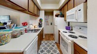 This single-story unit offers a practical and functional layout designed to maximize available space. The galley-style kitchen is efficiently organized, featuring wood-toned cabinetry lining both walls, white appliances including an electric range, microwave, dishwasher, and refrigerator, paired with light countertops and a single-basin sink. Hardwood-style flooring runs the length of the kitchen, adding warmth to the space.

The bedroom is comfortably sized with neutral carpet flooring and a textured ceiling. A window fitted with horizontal blinds and grommet-style curtain panels allows natural light to filter in while providing privacy. The room offers enough space to accommodate standard bedroom furnishings.

The bathroom continues the hardwood-style flooring found in other areas of the unit and features a wood-toned vanity with a light countertop, a toilet, and a tub-shower combination enclosed by a decorative curtain. Louvered cabinet doors add a subtle design detail to the space.

Throughout the unit, neutral wall tones create a cohesive, understated aesthetic, providing a versatile backdrop adaptable to various furnishing styles.