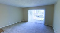 This single-floor residential space offers a clean, straightforward layout designed for comfortable everyday living. The living area is a standout feature, showcasing wall-to-wall carpeting in a neutral tone that adds warmth underfoot. A large sliding glass door, dressed with vertical blinds, serves as the focal point of the room, allowing an abundance of natural light to pour in while framing a pleasant view of the surrounding greenery and exterior surroundings. The neutral beige walls complement the soft carpeting, creating a calm and cohesive color palette throughout. The textured ceiling adds a subtle architectural detail that is consistent with the overall understated aesthetic of the space. Baseboards run cleanly along the perimeter of the room, providing a finished look. The open, uncluttered floor plan allows for flexible furniture arrangement, making it easy to personalize the living space to suit a variety of needs and styles. Overall, the space presents a functional and inviting environment, characterized by its bright, airy atmosphere and connection to the outdoors through its generous glazed access point.