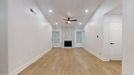 This single-floor unit showcases a clean, modern aesthetic unified by warm-toned wood-style flooring and crisp white walls throughout. The spacious living room is a standout, featuring soaring ceilings, recessed lighting, a ceiling fan, and a floor-to-ceiling marble-tiled fireplace flanked by two windows that invite natural light. A dedicated dining area is highlighted by an elegant multi-light pendant fixture, with open doorways connecting seamlessly to adjacent spaces. The kitchen is well-appointed with gray shaker-style cabinetry accented by gold hardware, white quartz countertops, a geometric patterned tile backsplash, and stainless steel appliances including a slide-in range with overhead microwave. Two bathrooms reflect the same contemporary design language: one features a tub-shower combination with large-format marble-look tile surround, while the other includes a vanity with matching cabinetry, gold and black hardware accents, and wood-look flooring. An additional room offers a versatile, open space suitable for a bedroom or flex use. The overall layout, as shown in the dollhouse view, is efficiently organized with a cohesive, freshly updated design throughout.
