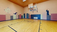 This indoor basketball court is a well-appointed athletic facility designed for serious recreational use. The court features a classic hardwood floor with clearly marked boundary and lane lines, providing a regulation playing surface suitable for basketball and other court sports. Soaring ceilings create an open, airy atmosphere and ensure ample vertical clearance for gameplay.

A wall-mounted glass backboard with a standard regulation hoop is positioned on one end of the court, extending from a sturdy steel bracket system anchored securely to the block wall. Along the perimeter walls, padded safety panels in a multicolored arrangement of blue, pink, green, and gray offer important player protection. These wall pads run continuously at lower wall height, adding both safety and visual character to the space.

The court's interior walls are constructed of painted concrete block and are accented by bold, athletic silhouette murals depicting players in action, reinforcing the sporting atmosphere. Fluorescent lighting overhead ensures bright, even illumination throughout the court.

On one side of the space, an interior window overlooks an adjacent fitness room equipped with exercise equipment, while glass doors nearby provide access and allow natural light to enter. The facility is well-ventilated, with HVAC vents integrated along the lower walls.