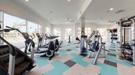 Located at 1902 Millenia Ave in Chula Vista, CA, this well-appointed fitness center offers a spacious, single-floor layout designed to support a comprehensive workout experience. The facility is flooded with natural light through large windows that line the perimeter walls, providing open views of the surrounding neighborhood and creating an energizing atmosphere throughout the space.

The main workout area is generously sized and houses a wide array of cardio equipment, including treadmills, elliptical machines, and stationary bikes, all thoughtfully arranged to maximize usability. A dedicated strength training zone features cable machines, weight benches, a full dumbbell rack, and additional resistance equipment. Floor-to-ceiling mirrors run along one wall, amplifying the sense of space and allowing users to monitor their form during exercise.

A large flat-screen television is mounted prominently on one wall, adding an entertainment element to workouts. Ceiling fans provide additional ventilation and comfort. The distinctive patterned carpet tiles in teal, beige, and dark brown add a modern, lively aesthetic to the interior. A separate, enclosed room off the main floor appears to serve as a secondary activity or studio space. Access to the facility is through glass-paneled double doors that open to a bright outdoor courtyard area, contributing to the seamless indoor-outdoor connectivity of the overall property.