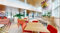Located at 222 N City Dr in San Marcos, CA, this striking commercial space delivers a dynamic and contemporary environment designed to inspire and engage. The open floor plan is anchored by soaring double-height ceilings in the main lounge area, where floor-to-ceiling windows flood the interior with abundant natural light and frame sweeping views of the surrounding cityscape. A bold, hand-illustrated mural wallcovering stretches across key walls, complemented by a curated collection of vibrant, colorful art panels that lend the space an energetic, creative character. The lounge area features a thoughtfully arranged mix of seating, including rounded sculptural armchairs in warm golden tones, a deep red sectional sofa, and chartreuse bench seating, all set atop a richly patterned area rug. A dedicated dining area offers a large oval table surrounded by coral upholstered chairs beneath a cascading sculptural pendant light, with a curved built-in counter and additional mural artwork nearby. Warm wood-panel ceilings, herringbone-patterned tile flooring, and glass-railed staircases add architectural refinement throughout. Lush indoor planters and greenery enhance the biophilic design, while the entry features double glass doors opening to an outdoor terrace. This space combines artistic vibrancy with functional elegance.