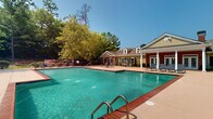 Located at 1400 Boxwood Blvd in Columbus, Georgia, this single-level outdoor amenity space centers around a generously sized swimming pool with clear, turquoise water and brick-accented coping. The pool area is well-appointed with multiple lounge chairs and seating areas arranged across a wide, tan-finished concrete sundeck, offering ample space for relaxation and recreation. A children's splash pad, featuring a distinctive red mushroom-style water feature, is situated adjacent to the main pool, making the area suitable for all ages. The perimeter of the space is enclosed by an iron fence, with manicured landscaping including ornamental shrubs, flowering crepe myrtles, and mature trees that provide shade and a sense of privacy. At one end of the pool, a classic Southern-style clubhouse commands attention with its yellow siding, red brick accents, white columned portico, and French doors that open directly onto the pool deck. The building is framed by lush greenery and flowering plants, enhancing its inviting curb appeal. The surrounding natural backdrop of rolling hills and tall pines adds a serene, park-like atmosphere to the entire outdoor environment. Additional seating and patio furniture are scattered throughout the deck, completing this well-designed community gathering space.