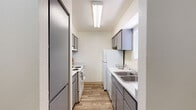 Located at 3535 N First Ave in Tucson, Arizona, this single-floor residence offers a well-organized layout designed for comfortable everyday living. The galley-style kitchen is efficiently arranged with gray cabinetry lining both walls, light speckled countertops, a double-basin sink, and white appliances including a range with coil burners, a dishwasher, and a refrigerator. Overhead fluorescent lighting and a window with blinds keep the kitchen bright and functional. Adjacent to the kitchen, the dining area accommodates a round glass-top table beneath a ceiling fan with integrated lighting, with a built-in pass-through shelf connecting to the kitchen. The living area features wood-look flooring, a textured ceiling, and a large sliding glass door dressed with vertical blinds that allows abundant natural light and outdoor access. The two bedrooms are carpeted and each includes a closet and windows with blinds for privacy. Both bathrooms are finished with gray vanity cabinetry, speckled countertops, wood-look flooring, and shower-tub combinations. A second bathroom features a separate vanity area leading to an enclosed shower. The overall aesthetic is clean and neutral, with consistent gray and white tones throughout the space.