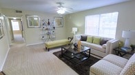 This single-story space features a well-appointed living area designed with comfort and style in mind. The room is finished with light-toned carpeting that extends throughout, contributing to a warm and cohesive atmosphere. A ceiling fan with an integrated light fixture is centrally positioned overhead, providing both air circulation and ambient lighting.

Large windows fitted with white horizontal blinds allow generous amounts of natural light to enter the space, creating an airy, open feel. The walls are painted in a soft, neutral tone that complements the overall design aesthetic.

The living area is thoughtfully arranged with a plush sofa accented by decorative pillows in earthy greens and grays, paired with a sleek black glass-top coffee table resting on a rich, dark area rug. A yellow upholstered bench adds a pop of color and serves as a functional accent piece. A slender metallic shelving unit displays decorative items and framed artwork, while coordinating wall art enhances the room's visual appeal. A floor lamp provides additional task lighting, rounding out a living space that balances practicality with contemporary design sensibility.