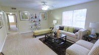 This single-story space features a well-appointed living area designed with comfort and style in mind. The room is finished with light-toned carpeting that extends throughout, contributing to a warm and cohesive atmosphere. A ceiling fan with an integrated light fixture is centrally positioned overhead, providing both air circulation and ambient lighting.

Large windows fitted with white horizontal blinds allow generous amounts of natural light to enter the space, creating an airy, open feel. The walls are painted in a soft, neutral tone that complements the overall design aesthetic.

The living area is thoughtfully arranged with a plush sofa accented by decorative pillows in earthy greens and grays, paired with a sleek black glass-top coffee table resting on a rich, dark area rug. A yellow upholstered bench adds a pop of color and serves as a functional accent piece. A slender metallic shelving unit displays decorative items and framed artwork, while coordinating wall art enhances the room's visual appeal. A floor lamp provides additional task lighting, rounding out a living space that balances practicality with contemporary design sensibility.