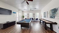 This single-floor community game room offers a well-designed recreational space ideal for socializing and leisure. The room is anchored by a full-size pool table with a blue felt surface, positioned at the center of the open floor plan, complemented by a shuffleboard table along one wall. Rich, dark hardwood-style flooring runs throughout, providing a warm contrast to the crisp white walls and textured ceiling.

Natural light flows generously through multiple windows and glass-paneled exterior doors, creating a bright and welcoming atmosphere. Dual ceiling fans maintain comfortable airflow across the space. A wall-mounted flat-screen television is positioned for easy viewing from multiple areas, accompanied by a media console below.

A casual seating area with bistro-style tables and chairs occupies one corner, offering a spot for conversation or tabletop games. Tasteful artwork and framed prints adorn the walls, adding a polished aesthetic. A large mirror near the entryway enhances the sense of openness, while potted greenery introduces natural accents throughout.

The entry area features clean, minimalist design with a wooden bench and direct access to adjoining spaces. Overall, this game room combines a lively, activity-focused layout with a clean, modern aesthetic, making it an inviting hub for community recreation.