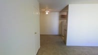This single-floor unit offers a straightforward, functional layout with a connected living and kitchen area. The living space is finished with wall-to-wall carpeting in a neutral tone, providing a comfortable underfoot feel throughout. A ceiling fan with a built-in light fixture is centrally mounted on the textured ceiling, offering both ventilation and ambient lighting for the room. The walls are painted in a clean, off-white tone, contributing to a bright and open feel.

Accessible directly from the living area, the kitchen is partially open to the main space, allowing for a connected flow between the two areas. The kitchen features wooden upper cabinetry, a refrigerator, and additional appliances, providing essential functionality. The transition from carpet to hard flooring marks the shift into the kitchen zone.

A doorway visible within the living area provides access to additional parts of the unit, suggesting further rooms or storage beyond what is seen. The textured ceiling runs throughout, consistent with the overall design. The space presents a clean, move-in ready environment suited for practical, everyday living.