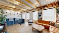 Sky Lounge is a well-appointed communal retreat that seamlessly blends warmth and modern design across a single floor. The space is immediately distinguished by its striking exposed wood beam ceilings set against a textured acoustic ceiling, lending a refined yet relaxed character throughout. Light wood-toned tile flooring flows continuously, unifying each area with a cohesive aesthetic.

The living room serves as the heart of the lounge, centered around a plush leather sectional sofa and a solid wood round coffee table. Floor-to-ceiling built-in shelving frames the room, displaying books, plants, and curated décor, while large windows with wood trim flood the space with diffused natural light. A wall-mounted flat-screen TV and additional seating complete this inviting gathering area.

The kitchen is equally impressive, featuring sleek grey cabinetry, dark stone countertops, and white textured tile backsplash. A standout design element is the collection of hanging potted plants suspended from above, adding a lively, organic touch. The kitchen is well-equipped with a sink, bar-height seating, and ample counter space, making it ideal for entertaining.

A patio extends the lounge outdoors, offering an additional venue for relaxation. Together, these spaces create a versatile, stylish environment perfect for socializing or unwinding.