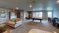 Located at 1606 Prairie Pl in Holmen, Wisconsin, this single-floor community clubhouse — known as Grasslands — is thoughtfully designed to offer residents a welcoming and well-appointed shared space. The grand entrance lobby immediately impresses with polished marble-look tile flooring, decorative wainscoting, and a striking octagonal tray ceiling with rich wood trim and an elegant chandelier. A sculptural metal Grasslands logo anchors the main corridor, which also features a bank of resident mailboxes. Adjacent to the entry, a comfortable lounge area invites relaxation with a gas fireplace, plush seating, and a wall-mounted television — an ideal spot to unwind. The full community kitchen is a standout, showcasing rich cherry cabinetry, granite countertops, stainless steel appliances including a refrigerator, range, and microwave, and a large center island with seating. A dedicated dining area with dark wood tables and a mounted flat-screen television provides a communal gathering spot. The game room offers generous open floor space, a full-size pool table, and natural light through large windows. A private office, furnished with a desk, shelving, and filing storage, rounds out the amenities, providing a quiet workspace for residents.