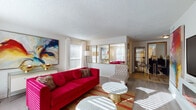 This single-floor residence makes a striking visual statement through its bold, curated interior design. The living room serves as the heart of the space, featuring light hardwood-style flooring, a textured ceiling, and abundant natural light streaming through large windows dressed in sheer white curtains. A built-in media wall with an electric fireplace anchors the room, flanked by vivid large-scale abstract artwork that adds color and energy. The open layout flows naturally toward a dining area, connected by a mirrored partition that enhances the sense of depth and light.

The kitchen is clean and functional, outfitted with white cabinetry, dark countertops, a tile backsplash, and stainless hardware, providing ample storage and workspace.

Both bedrooms are generously sized with plush carpeting and large windows that invite natural light. One bedroom features a bold red checkered headboard and a gallery wall of round mirrors, while the second showcases a red and white color scheme with statement artwork above the bed. Gold-accented arched mirrors and refined furnishings lend an elegant touch throughout.

A dedicated vanity area connects the bedroom wing, complete with gold-framed mirrors and stylish seating, rounding out this cohesive and design-forward residence.