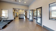 This single-story fitness center offers a thoughtfully designed space dedicated to health and wellness. The layout is open and spacious, providing ample room to move between a variety of cardio equipment, including a row of treadmills and elliptical machines, each equipped with individual digital displays for an enhanced workout experience. The flooring throughout is a durable wood-look material that adds a clean, modern aesthetic while remaining practical for high-traffic use.

A standout architectural feature is the prominent arched window grouping along one wall, which floods the cardio area with natural light and offers views of the surrounding outdoor greenery—creating an inviting atmosphere that encourages regular use. Additional windows positioned along adjacent walls further enhance the brightness of the space.

Recessed lighting is installed throughout the ceiling, ensuring consistent, even illumination during early morning or evening sessions. The entry area includes a corridor with built-in shelving, providing convenient storage for personal items. Glass-panel double doors connect this entry zone to the main workout floor, contributing to a sense of openness and transparency throughout the facility. Neutral wall tones and clean architectural lines give the space a polished, professional appearance.