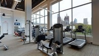 This contemporary fitness center is defined by its striking floor-to-ceiling glass window wall, which floods the space with abundant natural light and frames an impressive urban skyline backdrop. The expansive glazing creates an open, airy atmosphere that makes workouts feel energizing and connected to the city beyond.

The facility is well-equipped with a comprehensive selection of commercial-grade exercise machines, including multiple treadmills positioned to take full advantage of the panoramic views, as well as a variety of strength-training and cable machines suited for full-body workouts. A dedicated free weights and functional training area is also visible, catering to a range of fitness preferences and routines.

The interior finishes reflect a modern, industrial aesthetic, with exposed ceiling elements and neutral-toned carpeting that complement the sleek, dark-framed equipment. Pendant lighting fixtures supplement the generous natural light, ensuring the space is well-lit at any hour. The layout is thoughtfully organized to allow comfortable circulation between equipment stations. Overall, the fitness center offers a polished, professional environment with an exceptional urban outlook.