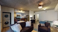 This single-floor studio offers a thoughtfully designed open-concept layout that seamlessly integrates the living, dining, sleeping, and kitchen areas into one cohesive space. A defining architectural feature is the exposed concrete ceiling, which runs throughout and lends an industrial-chic character complemented by warm lighting from ceiling fans and track lighting.

The kitchen is well-appointed with stainless steel appliances, including a range, microwave, refrigerator, and dishwasher. Rich wood-toned lower cabinetry is paired with white upper cabinets and dark countertops, providing ample storage and prep space. A stacked washer and dryer unit is conveniently tucked within the kitchen area, adding practical in-unit laundry access.

The dining area features a compact round table with modern chairs, positioned adjacent to the kitchen for easy functionality. The open living area accommodates a comfortable seating arrangement and flows directly into the sleeping zone, which is defined by a full-size bed framed by exposed concrete accents and neutral walls.

Light wood-tone flooring runs throughout the main living space, enhancing the bright, airy feel of the interior. The bathroom features a contrasting dark terrazzo-style floor, a wall-mounted storage cabinet, and a shower-tub combination, completing this well-rounded urban residence.