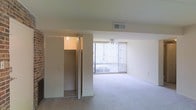 This single-floor space features an open living area finished with wall-to-wall carpeting that lends a soft, neutral aesthetic throughout. A large sliding glass door dominates one wall, fitted with vertical blinds that allow for adjustable natural light and provide access to an outdoor area. A ceiling fan is centrally positioned overhead, offering both ventilation and subtle visual interest to the room.

The living area is generously proportioned, providing ample space for a variety of furniture arrangements. The walls are finished in a clean, neutral tone that complements the light carpeting and contributes to the overall bright and airy feel of the space.

Adjacent to the main living area is a walk-in closet, visible through open bifold doors. The closet is equipped with built-in shelving and hanging storage, offering practical organizational capacity. A ceiling light fixture within the closet ensures visibility and ease of use.

An overhead HVAC vent is integrated into the ceiling, supporting climate control throughout the space. The layout is straightforward and functional, with clean lines and minimal architectural ornamentation, making it well-suited for a range of furnishing styles.