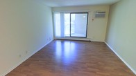 This single-floor space offers a straightforward and functional layout designed for comfortable everyday living. The main living area is highlighted by rich hardwood-style flooring that runs throughout, lending warmth and a polished look to the interior. Neutral, light-toned walls create a bright and airy atmosphere, complemented by a textured ceiling that adds subtle architectural character.

A large sliding glass door serves as the focal point of the living area, flooding the space with natural light and providing direct access to an outdoor balcony. Vertical blinds offer flexible light control and privacy. The balcony overlooks the surrounding exterior, offering a pleasant outdoor retreat. A wall-mounted air conditioning unit ensures climate control, while a baseboard heating system provides additional comfort during cooler months.

The overall ambiance of the space is clean, open, and understated, making it well-suited for a variety of furnishing styles. The combination of natural light, functional climate systems, and durable flooring makes this a practical and inviting space for everyday use.