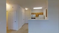 This single-floor space is organized around a functional, open-concept kitchen and hallway layout. The kitchen is equipped with warm honey-toned wood upper cabinets that provide ample storage, complemented by white appliances including a refrigerator, microwave, and range. A white peninsula counter extends into the living area, creating a natural division between the kitchen and the adjoining space while also offering additional prep or casual dining surface. Overhead fluorescent lighting illuminates the kitchen workspace effectively. The flooring throughout is light-toned tile, lending a clean, cohesive look to the interior. Crown molding details along the ceiling add a subtle architectural refinement to the otherwise straightforward design. A hallway extends from the main area, lined with multiple white panel doors that lead to additional rooms, suggesting a practical and well-divided floor plan. The walls are painted in a neutral white throughout, keeping the space feeling bright and open. The overall ambiance is clean, utilitarian, and move-in ready, with a layout designed to maximize usability across the available footprint.