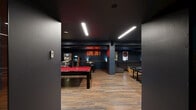 Located at 1090 Rue De Bleury in Montréal, this expansive games room is a meticulously designed entertainment hub that seamlessly blends bold aesthetics with a wide range of recreational activities. The space is finished with warm hardwood flooring throughout, set against deep navy and charcoal walls that create an immersive, lounge-like atmosphere enhanced by sleek linear LED ceiling lighting.

At the heart of the room sits a striking red-felt pool table, flanked by a full-size ping pong table. A dedicated arcade alcove — accented by exposed brick walls and a glowing red neon "GAME ON!" sign — houses classic pinball machines and arcade cabinets alongside a foosball table, giving the space a vibrant retro energy.

A projector and large projection screen anchor a screening area, complemented by iconic white and red ball chairs that serve as both statement pieces and comfortable seating. A generously sized sectional sofa lounge, decorated with framed artwork and natural wood stump side tables, provides a relaxed retreat for spectators and socializing.

A small kitchenette area with cabinetry and a sink offers added convenience. The overall design is cohesive and intentional — dark, dramatic, and purpose-built for entertainment, making this games room an exceptional amenity.