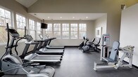 The fitness center at Townley Park is a well-equipped, single-floor facility designed to support a comprehensive workout routine. The space is characterized by its clean, modern aesthetic, with neutral-toned walls, recessed lighting, and durable rubber flooring throughout. Large multi-pane windows line the perimeter walls, allowing generous natural light to fill the interior while providing views of the surrounding outdoor landscape, creating an open and energizing atmosphere.

The cardio area is prominently featured and includes multiple treadmills, elliptical trainers, and stationary and recumbent bikes, all positioned to take advantage of the natural light from the windows. A wall-mounted television is suspended from the ceiling, offering entertainment during workouts. The varied ceiling height, with a raised section adding volume to part of the space, contributes to an airy feel throughout the room.

A dedicated strength training section houses a selection of selectorized weight machines, including leg press and cable stations, providing options for targeted muscle conditioning. The layout is thoughtfully arranged to allow clear circulation between equipment zones. Overall, the fitness center presents a functional, well-lit environment suited for a range of fitness activities.