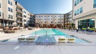 Located at 525 E 21st St in Charlotte's vibrant 28205 corridor, this outdoor amenity space is designed to deliver a resort-style experience within a modern residential community. At its centerpiece sits a generously sized swimming pool with crystal-clear blue water, flanked by a shallow wading entry and fountain features that add a playful touch to the aquatic environment. The pool deck is well-appointed with rows of contemporary lounge chairs featuring bold lime-green cushions, offering ample space for sunbathing and relaxation under the Carolina sky.

Surrounding the pool, the courtyard opens into distinct zones for socializing and leisure. A covered pergola area with string lighting provides a shaded retreat with seating arrangements and raised planters filled with seasonal flowers, creating a warm, inviting atmosphere. The hardscape throughout combines polished concrete and stone masonry detailing, contributing to the refined aesthetic of the outdoor space.

The courtyard is enclosed by a multi-story building with a curvilinear facade, balconies, and large-pane windows that overlook the amenity area. An indoor amenity room accessible via expansive floor-to-ceiling glass doors connects seamlessly to the outdoor deck, blurring the line between interior and exterior living. The overall design emphasizes open-air communal living with thoughtful landscaping and modern furnishings throughout.