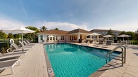 Located on Allure Circle in Orange City, Florida, this outdoor community amenity space offers a resort-style experience designed for relaxation and recreation. At its centerpiece is a generously sized swimming pool with crystal-clear blue water, framed by a broad, light-toned paver deck that surrounds the pool on all sides. The deck is well-appointed with rows of contemporary sling-style lounge chairs and shaded umbrella stations, providing ample space for sunbathing and poolside leisure. A covered pavilion anchors one end of the space, featuring a coffered ceiling with a ceiling fan for added comfort in Florida's warm climate. The pavilion is furnished with modern outdoor sofas and armchairs in neutral tones, creating an inviting social lounge area that opens directly to the pool deck. Adjacent to the pavilion, an outdoor kitchen or bar area provides additional amenity space for gatherings. The perimeter of the pool area is enclosed by a wrought-iron style fence and lush green hedging, offering both privacy and a polished aesthetic. Tall palm trees and tropical foliage throughout the grounds reinforce the relaxed, Florida-inspired atmosphere. The clubhouse structure features a hip roof with architectural shingles and a light exterior, complementing the overall bright and airy character of this well-designed outdoor community space.