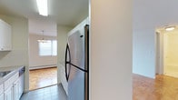 Located at 10 Landing Lane, this single-floor residence features a clean, functional layout with consistent parquet wood flooring throughout the living areas and bedrooms. The warm, geometric pattern of the parquet adds visual character and a sense of cohesion across the space.

The living area is generously proportioned, illuminated by a wide bank of windows that allow ample natural light to fill the room. A chandelier provides additional lighting, and a wall-mounted air conditioning unit and baseboard heating ensure year-round comfort. The open sightlines create an airy, welcoming atmosphere.

The kitchen is efficiently designed with white cabinetry, a tile backsplash, and stainless steel appliances including a refrigerator, electric range, over-the-range microwave, and a stainless steel sink. Tile flooring in the kitchen provides a practical transition from the parquet in the adjoining areas.

The bedroom offers a spacious feel with the same parquet flooring and features a built-in closet with bifold doors, providing organized shelving and hanging space. A window brings in natural light, and a wall unit offers supplemental climate control.

The bathroom is neatly appointed with a full tub and shower combination, white subway tile surround, a vanity with sink, and a mirrored medicine cabinet with built-in lighting above.