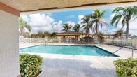 Located at 1720 NW Third Terrace in Florida City, Florida, this single-story community amenity facility is centered around a generously sized outdoor swimming pool that serves as the focal point of the space. The pool area is finished with a broad concrete deck ideal for lounging, complete with metal entry handrails and clearly marked depth indicators along the pool's edge. A white metal perimeter fence encloses the pool area, providing both safety and a defined boundary to the outdoor space.

The support building features a warm, stucco exterior painted in a distinctive golden-yellow hue with terracotta accent banding, lending the facility a welcoming, South Florida character. The building houses separate men's and women's restroom facilities, accessible via a covered walkway corridor. A wall-mounted water fountain is conveniently positioned between the restroom entrances, serving pool guests. The covered overhang provides a shaded transition space that connects the interior amenities to the pool deck.

A covered outdoor pavilion or gazebo structure with a dark shingled roof is visible beyond the pool fence, offering a shaded gathering spot. Mature tropical palm trees are scattered throughout the grounds, enhancing the lush, resort-like atmosphere. The facility benefits from abundant South Florida sunshine, creating a bright, open-air environment that is inviting year-round.