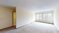 Located at 333 Oak Grove St in Minneapolis, MN, this one-bedroom, one-bathroom unit offers a functional and straightforward layout across a single floor. The living room is generously proportioned, featuring wall-to-wall carpeting, neutral cream-toned walls, and a large multi-pane window that brings in natural light and urban views of the surrounding neighborhood. A baseboard heating system runs along the perimeter, ensuring year-round comfort. The kitchen is efficiently designed with white cabinetry, countertop workspace, and standard appliances, positioned conveniently off the main living area near the entryway. The bedroom is a comfortable, well-sized room with carpeted flooring, its own large window overlooking neighboring buildings, a wall-mounted air conditioning unit, and baseboard heating. The neutral palette throughout keeps the space feeling bright and open. The bathroom is clean and classic, featuring white square tile surrounding a full soaking tub with an overhead shower, a vanity with sink, and mosaic tile flooring. A dark wood door frames the bathroom entrance, adding a touch of contrast. This unit provides a practical and livable space in the heart of Minneapolis.